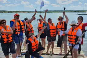 Eine überglückliche Gruppe auf eigenem Floß beim Floßbau Teamevent/Teambuilding/Betriebsausflug von bo events am Hardtsee zwischen Heidelberg und Bruchsal.